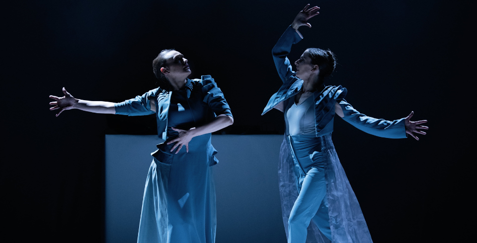Two dancers in a blue clothing with outstretched arms and open hands, both facing each other, creating a striking, symmetrical pose in dramatic lighting.