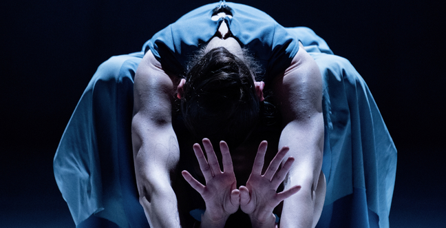 A dancer in a blue clothing bends forward with outstretched arms and open hands, creating a striking, symmetrical pose in dramatic lighting.