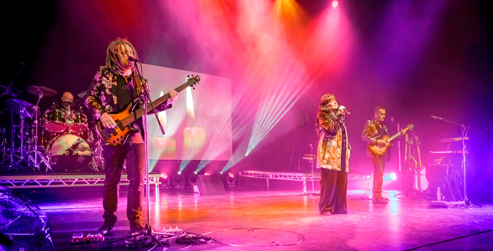 Lit by red and blue lights, two male guitarists and a female singer perform on stage