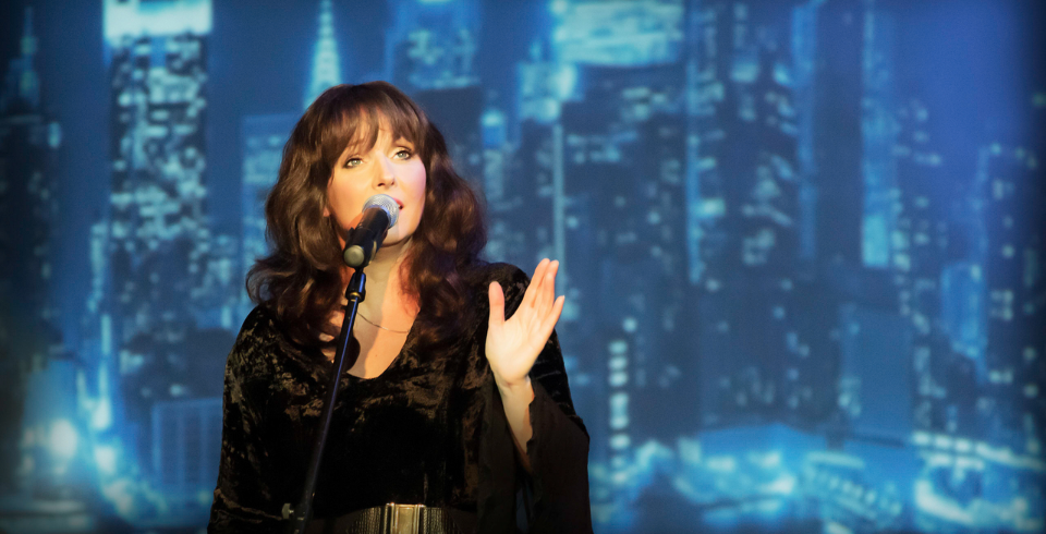 A brunette woman with a fringe sings into a microphone while stood in front of a projection of a cityscape
