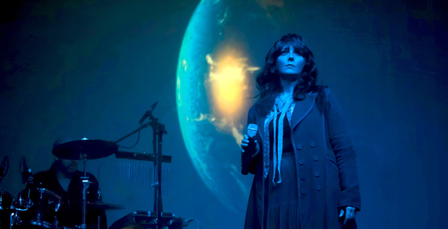 A woman with long brown hair with a fringe, wears a long coat and holds a microphone while standing in front of a projection of the Earth, lit in blue light