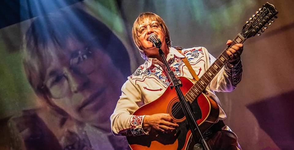 A man with long blonde hair in a country shirt with an acoustic guitar stands in front of a screen