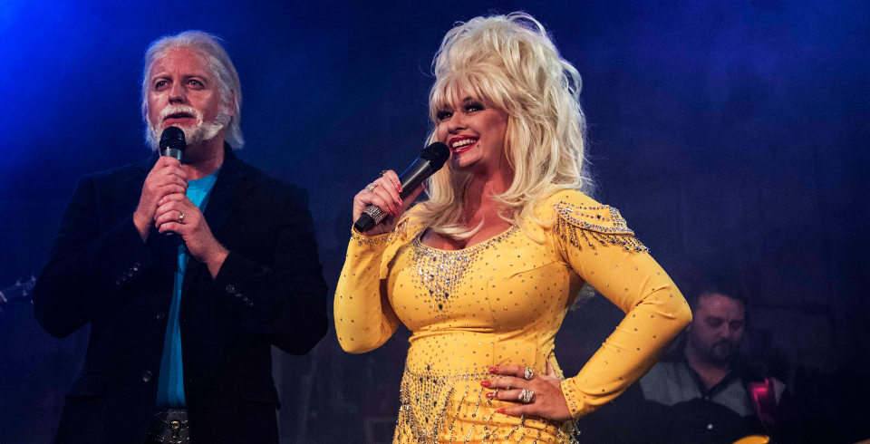A woman with blonde hair and a yellow glittery dress stands with her hand on her hip next to a man in a suit