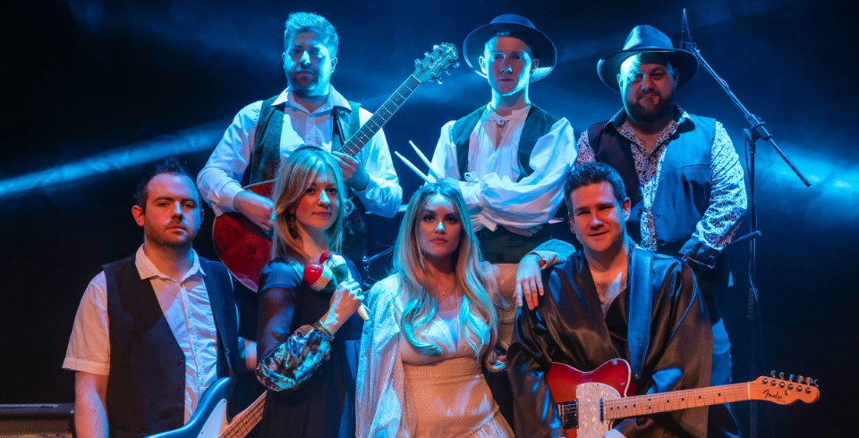 seven band members stand together. Three men at the back and two women with two men at the front. The men hold guitars.