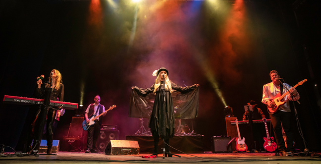 A band performs on stage with colorful lights and fog. The lead singer, dressed in a flowing black outfit and hat, stands in the center with arms outstretched. Other band members play keyboards, guitars, and drums.