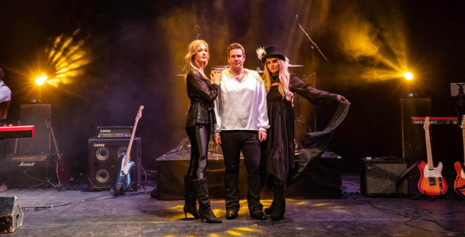Centre stage, one man with a woman either side of him. They are surrounded by guitars, amps amd other instruments. Stage lighting glares in the background.