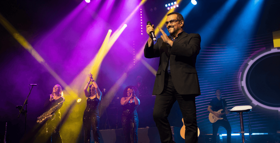 A singer in a black suit and sunglasses performs on stage with a live band, featuring backup singers and a saxophonist, under colorful stage lighting.