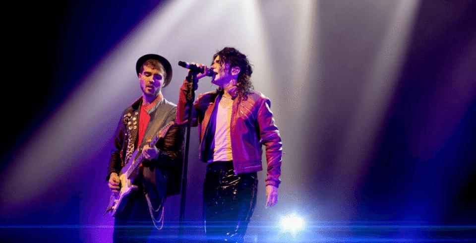 A man in a red leather jacket dressed as Michael Jackson stands by a guitarist on stage