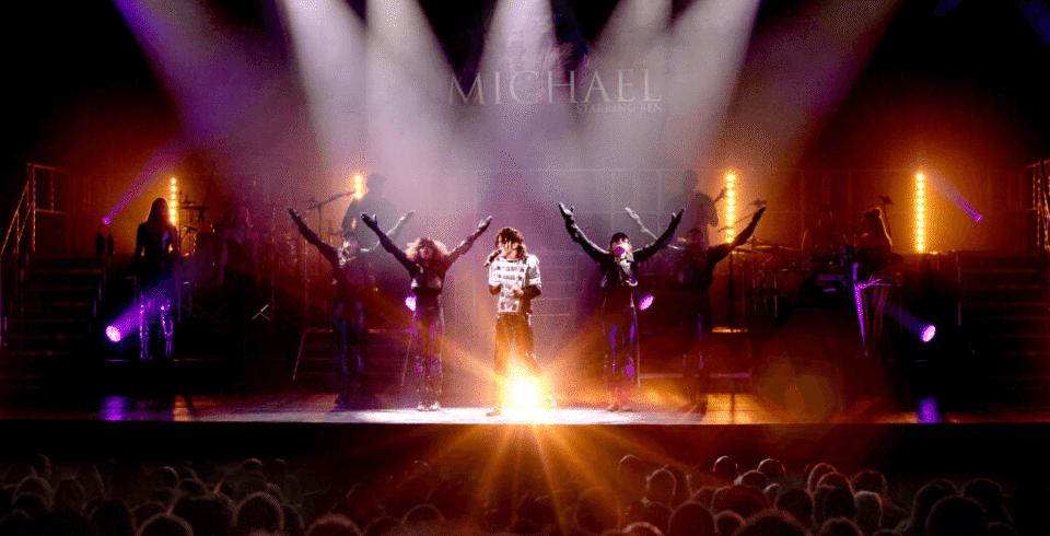 A group of dancers with their arms up on stage with spotlights facing down and warm yellow lighting and a man in the centre of the group dressed as Michael Jackson with a black hat and white top