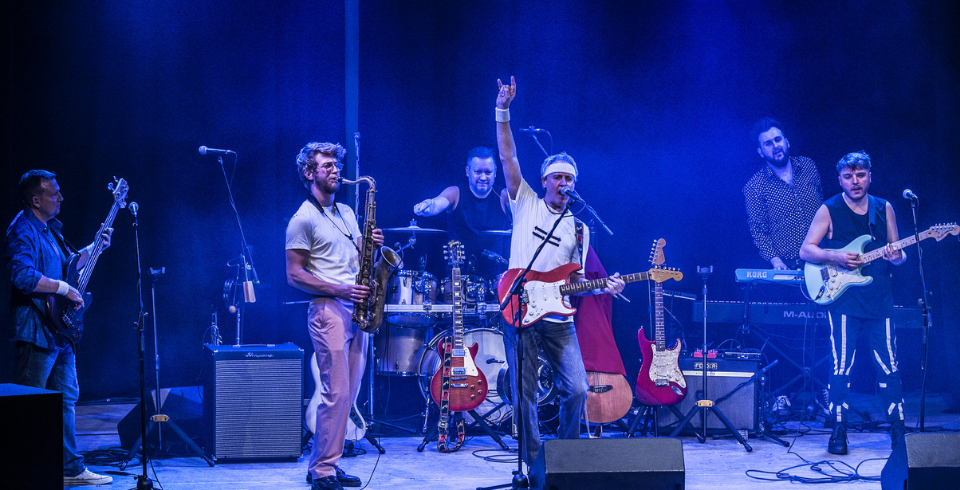A rock band performs live under blue lights, with the lead singer raising a hand while playing a red electric guitar.