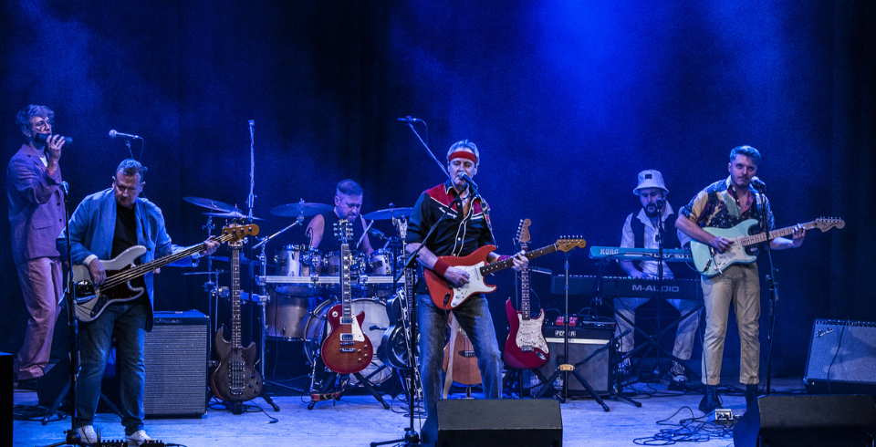 A rock band performs live under blue lights, the lead is playing a red electric guitar, with two other musicians either side of him also playing guitars.