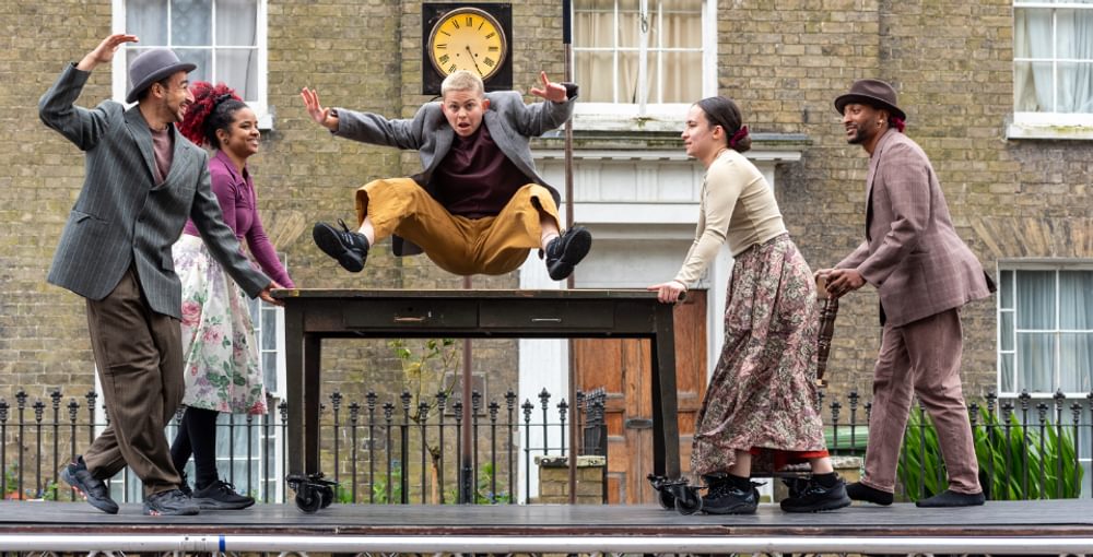 A performer jumps over a table which is being passed under them by 4 other performers, all on a giant treadmill
