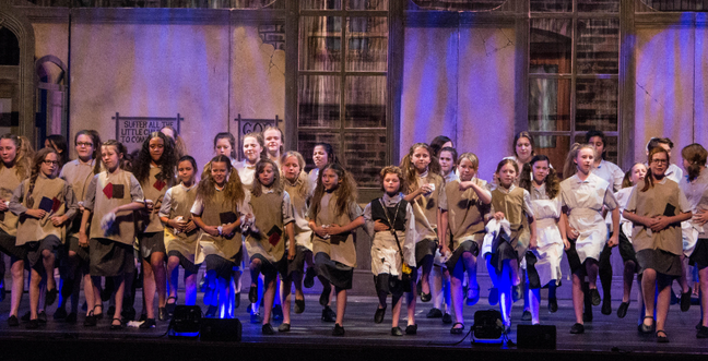 A group of children in a theatre production on stage wearing brown clothing in front of a room set with blue low-lights