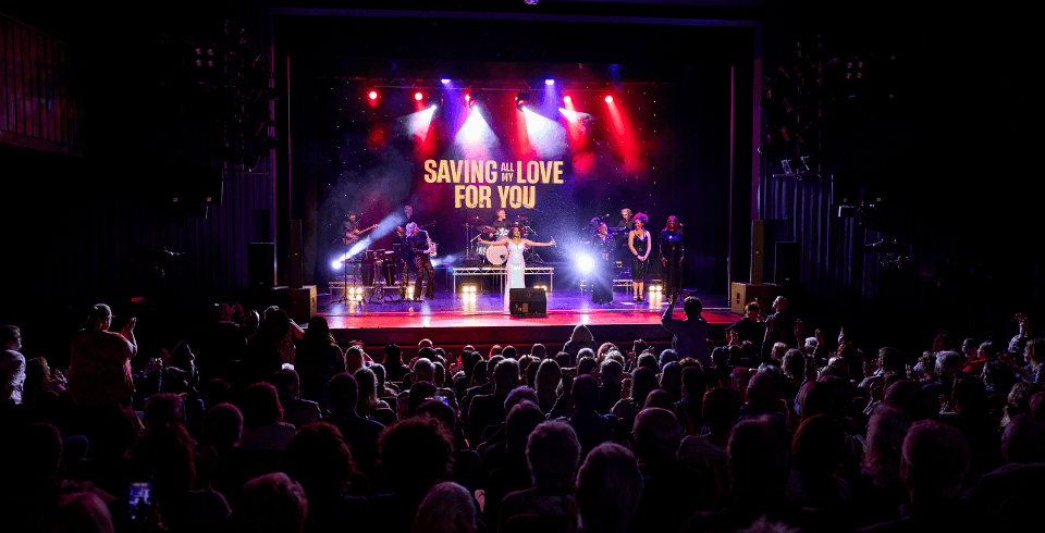 A woman stands on stage in a silver dress singing below the title 'Saving All My Love For You'