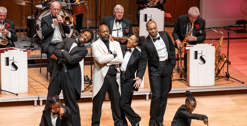 A jazz band performs on stage. In front of them, five dancers in black and white suits pose together. The stage is set with vintage-style white music stands labeled "HS."