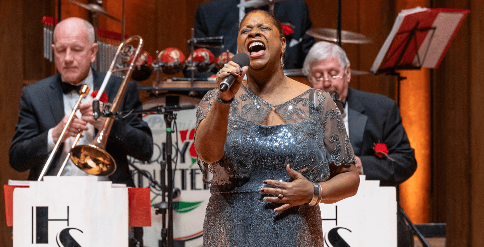 A powerful female vocalist in a sparkling silver dress passionately sings into a microphone, her hand on her stomach. Behind her, the jazz band performs.