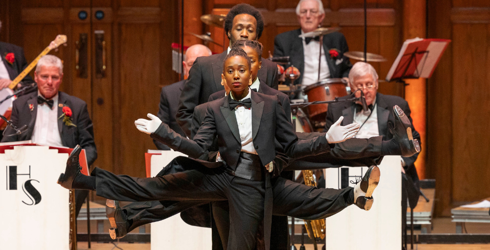 A striking choreographed dance moment where a female dancer in a tuxedo stands in the foreground with white gloves, while two dancers behind her extend their legs and arms outward in perfect symmetry. The jazz band provides musical accompaniment in the background.