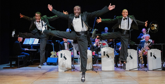 Three male tap dancers, dressed in sharp black tuxedos with white shirts and black suspenders, leap into the air in perfect unison.