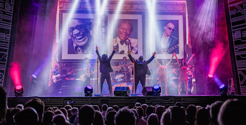 Two men dressed as the Blues Brothers on stage with their arms in the air