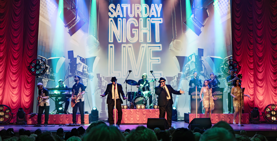Two men dressed as the Blues Brothers on stage in front of a sign that says 'Saturday Night Live'
