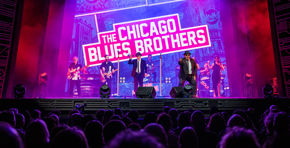 Two men dressed as the Blues Brothers on stage in front of a sign that says 'The Chicago Blues Brothers'