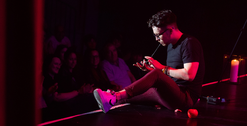 A man with short dark hair, glasses and a black t-shirt sits on the edge of the stage turned towards the audience