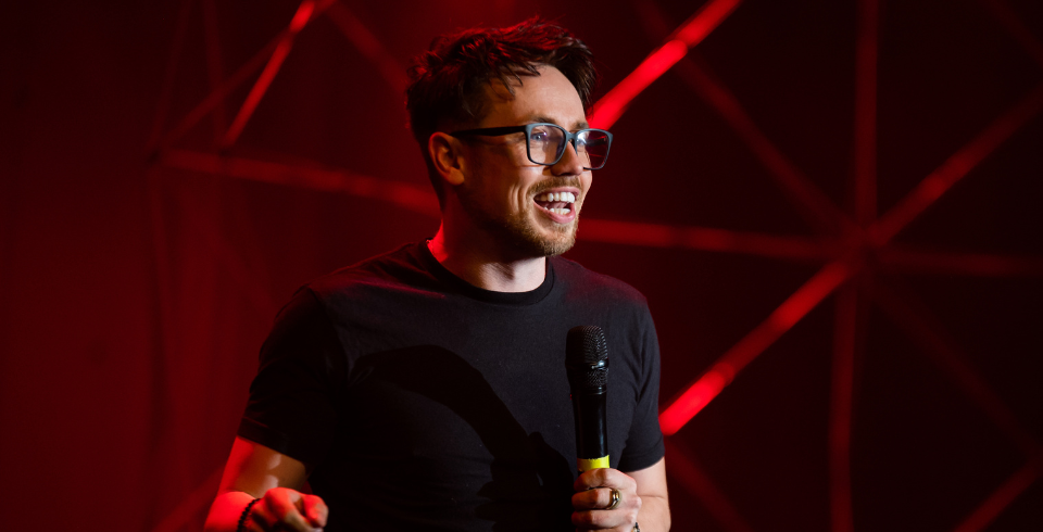 A man with short dark hair, glasses and a black t-shirt stands on stage holding a microphone. The background is lit with a red hue.