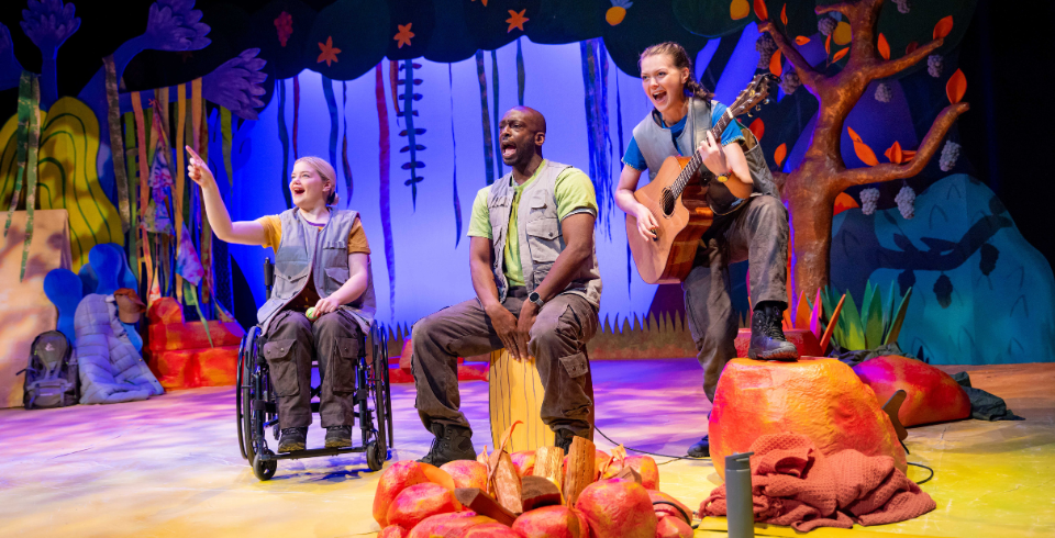 A group of people on a colourful stage sit around a campfire while one woman holds an acoustic guitar