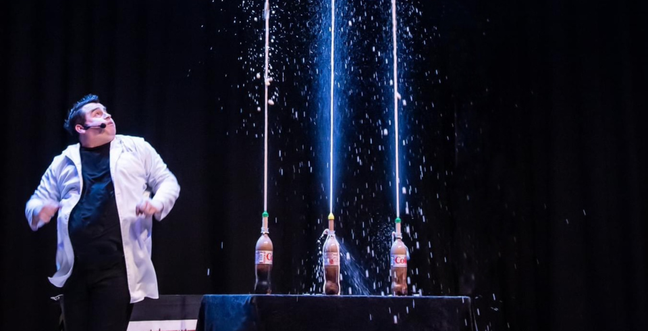 A male presenter in a lab coat reacts as three bottles of Diet Coke erupt in a synchronized Mentos experiment. The high-pressure soda streams shoot up into the air, creating an exciting visual.