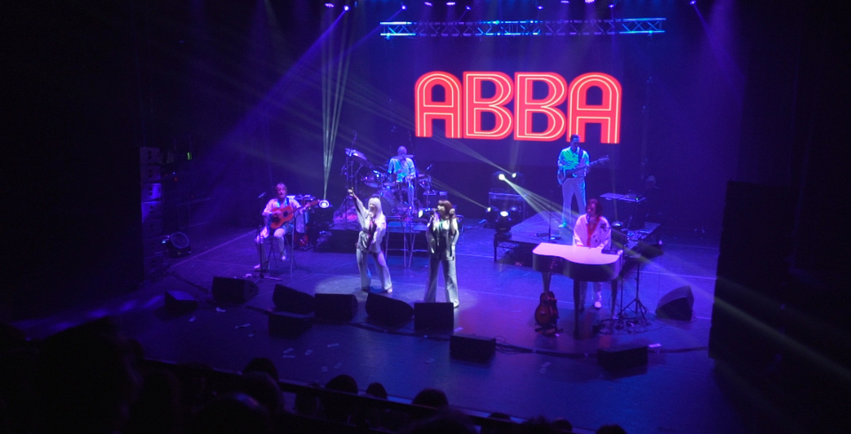 A band on stage with blue lights and the words 'ABBA' in red on stage behind them