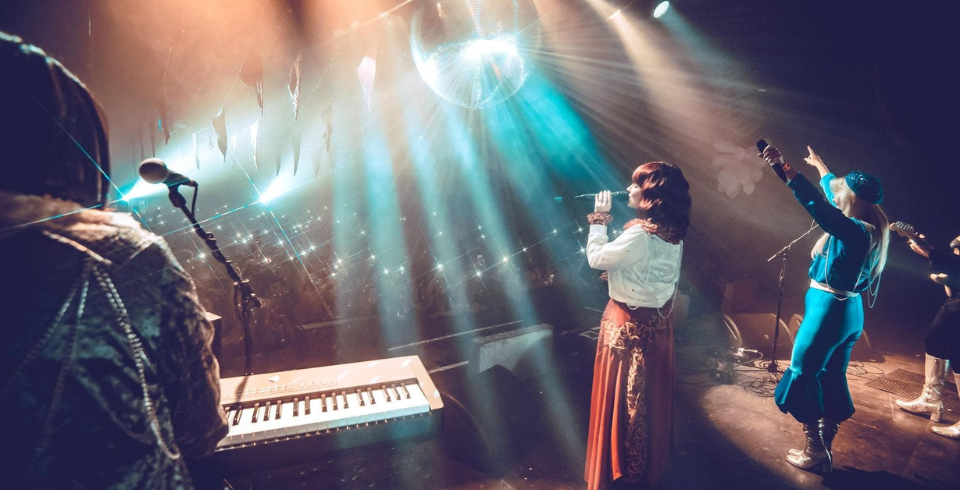 A band on stage with a keyboard, two women sing into microphones while blue lights shine on them