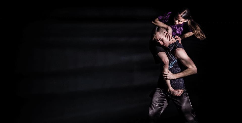 A male and female dancer perform an expressive movement. The male leans forward, while the female clings to his back, against a dark background.