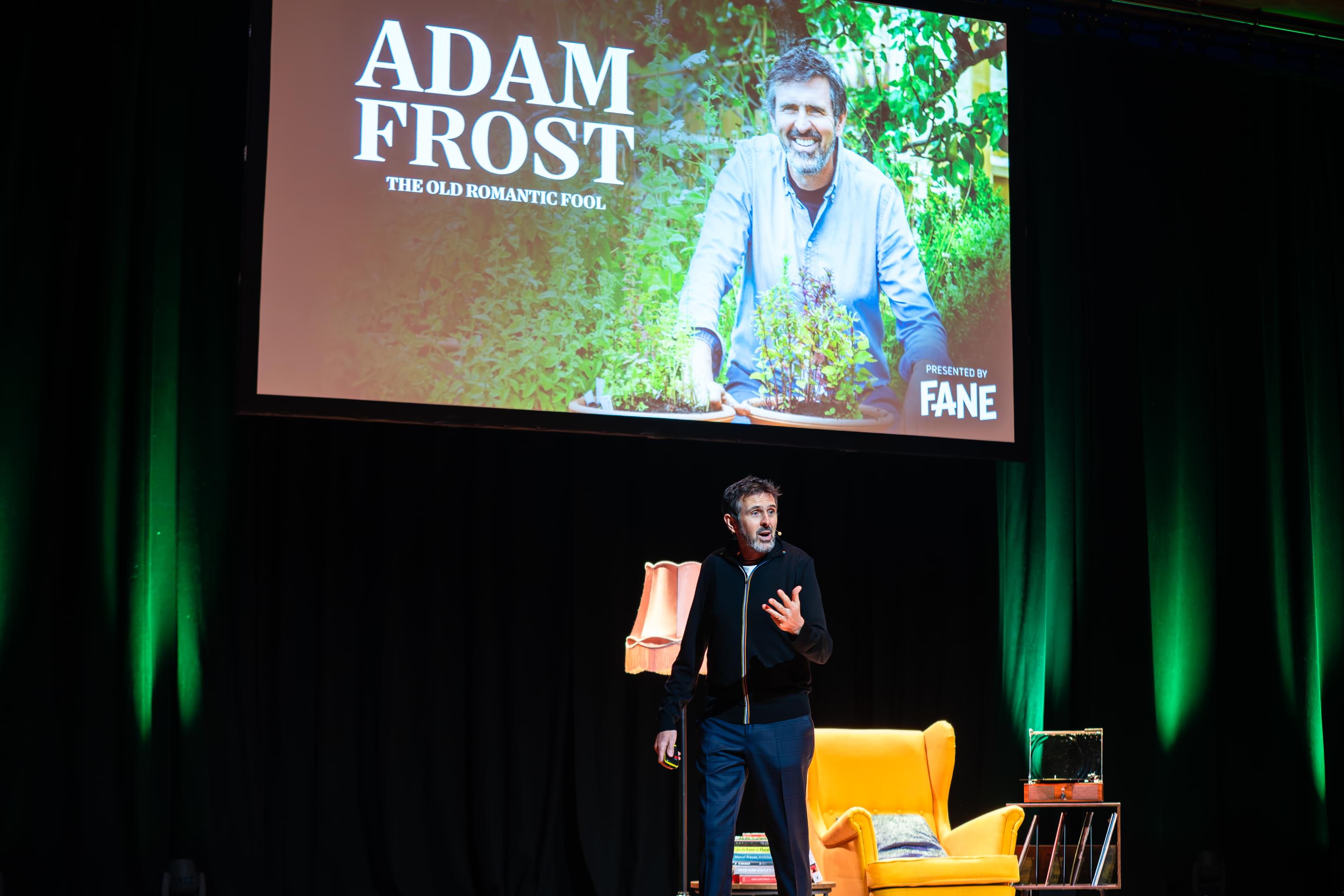 A man on a stage in front of a green curtain and a screen with 'Adam Frost' written on it with a yellow arm chair and lamp with a small microphone