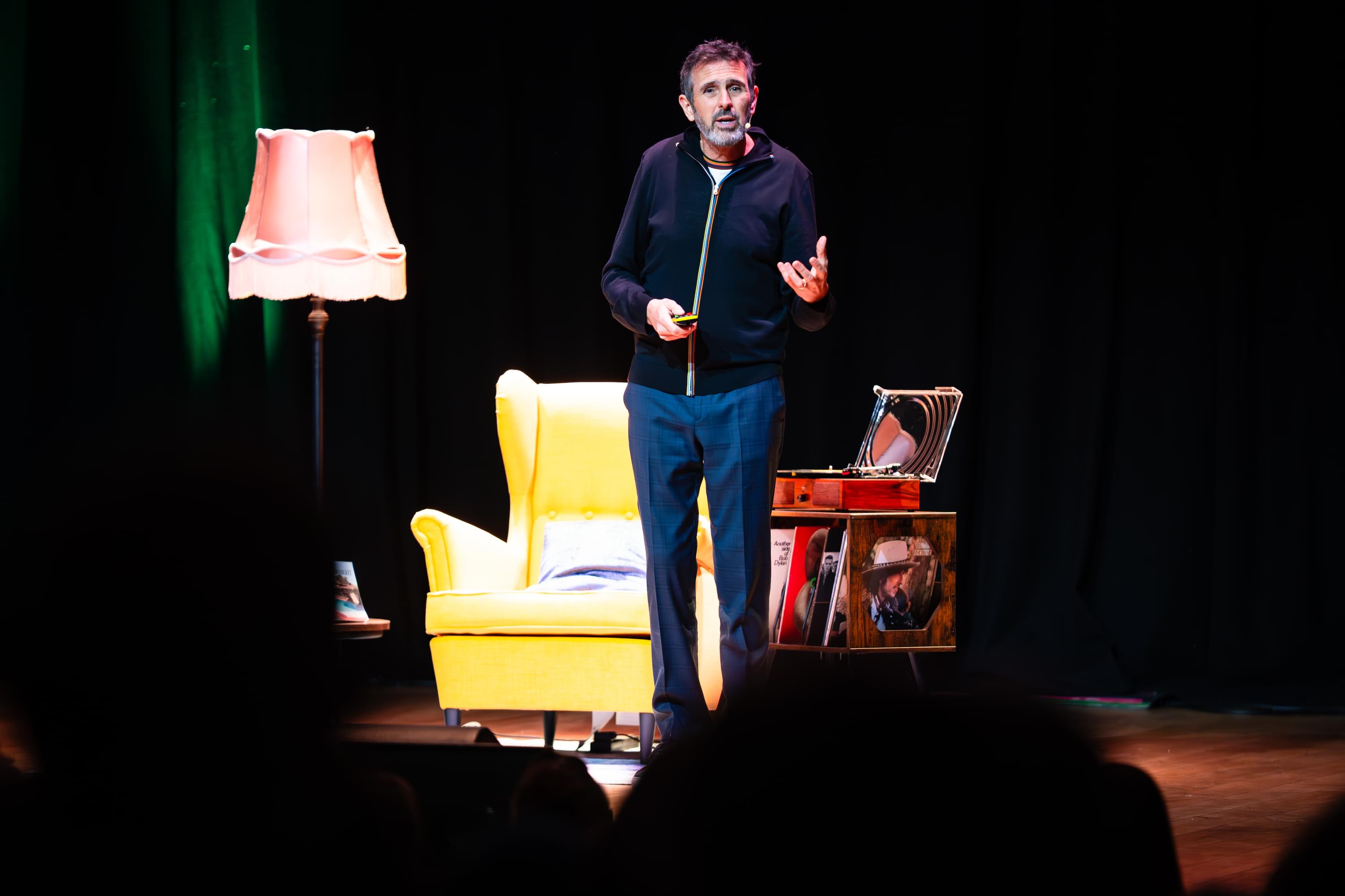 A man on a stage with a yellow arm chair and lamp with a small microphone