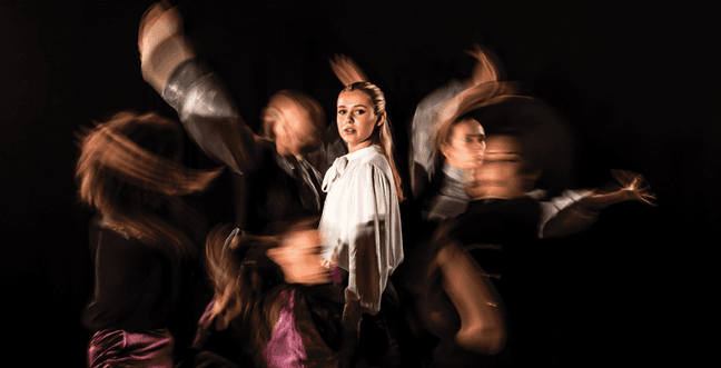 A girl in a white top in front of a group of dancers in a blur