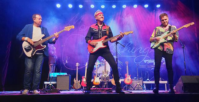 Three musicians all play their electric guitars together, backlit by blue and purple lights.