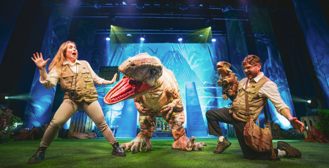 Two performers on stage interact with life-sized dinosaur puppets, one large and one small, in a theatrical jungle-themed setting.