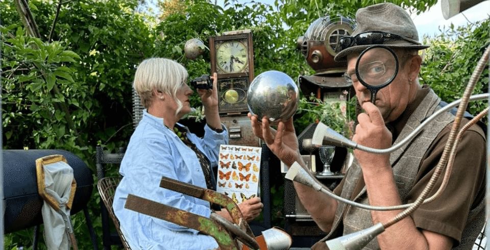 Two people surrounded by various objects, a man holds a magnifying glass and a silver ball and a woman looks into binoculars