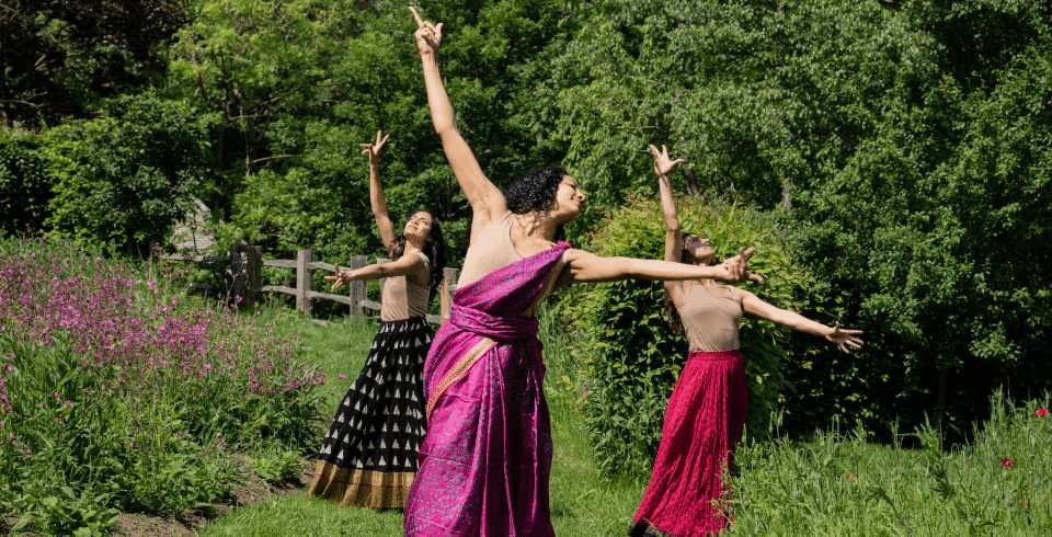 Three women with arms outstretched stand in formation, wearing saris in a green field of trees