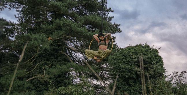 A woman hangs mid-air in trees on a hoop with her hand covering her face