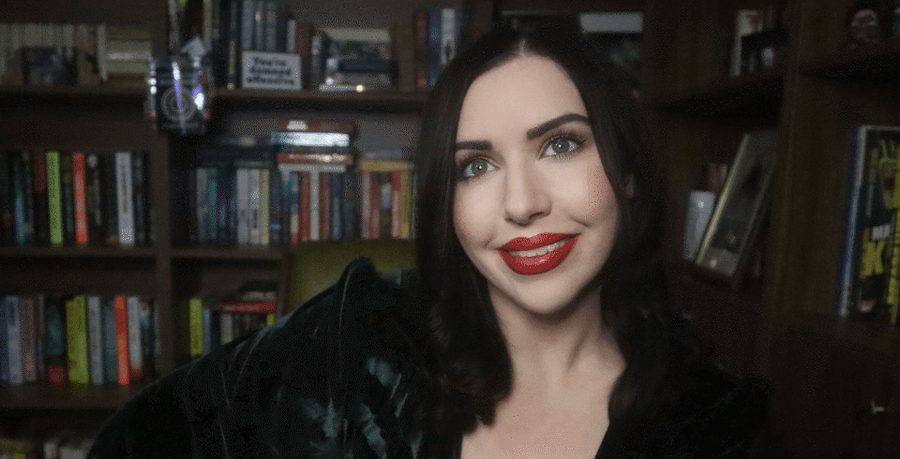 A woman wearing red lipstick and a black dress sits in front of a bookcase.