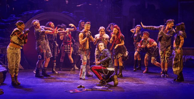 An ensemble of actors on a stage, all wearing punk-rock style clothes, surrounding a man who crouches down in the middle of them.