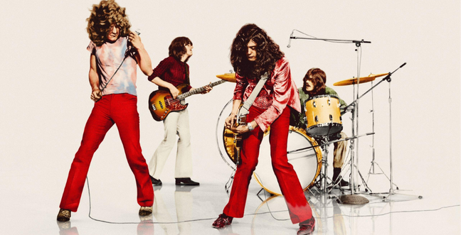 In a white room, a group of men play instruments. Two men at the front have red trousers and long hair.