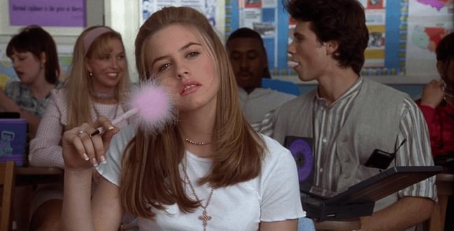 A girl with blonde hair and a white t-shirt sits in a class room holding a pink pen with a fluffy end to the pen