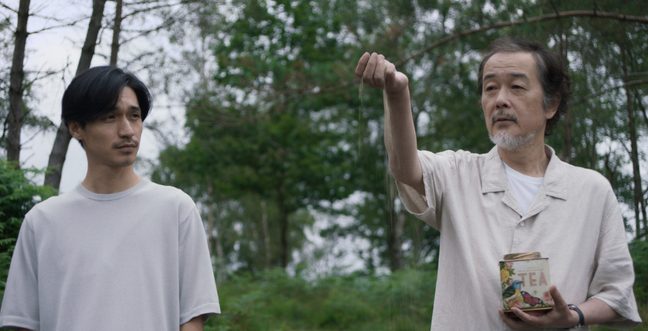 Two men in white tops stand in nature while one man holds up a jar of ashes