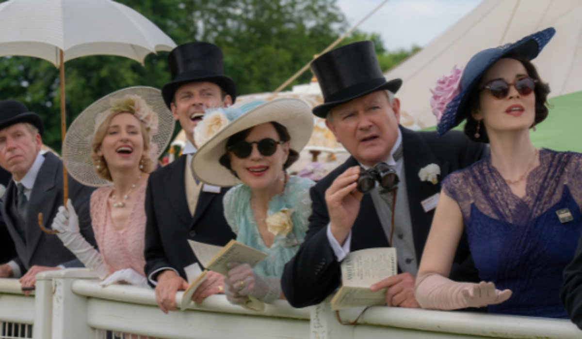 A group of elegantly dressed people standing by a railing. They are wearing vintage-style outfits with hats, gloves, and formal suits. The group looks excited and engaged, some holding binoculars and papers.