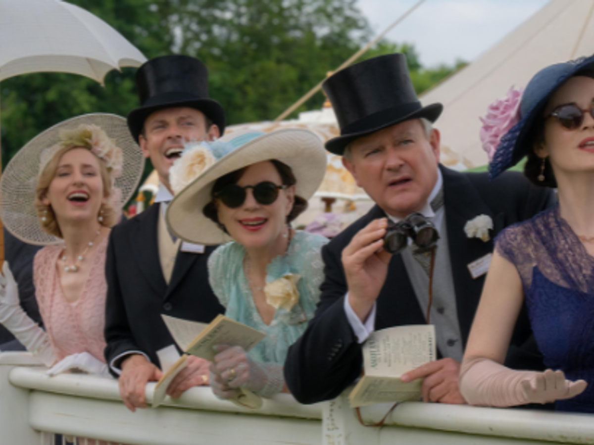 A group of elegantly dressed people standing by a railing. They are wearing vintage-style outfits with hats, gloves, and formal suits. The group looks excited and engaged, some holding binoculars and papers.