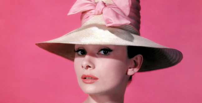 Audrey Hepburn stands against a pink background wearing a wide brimmed hat with a pink bow