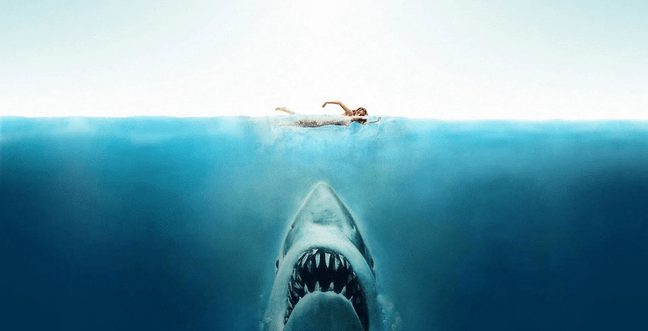 A person swims along the surface of the water as a shark comes up from below