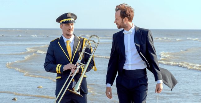 Two men walk along a beach, one in a sit with a white bow tie and the other in a suit with a yellow tie and hat, while holding a trumpet